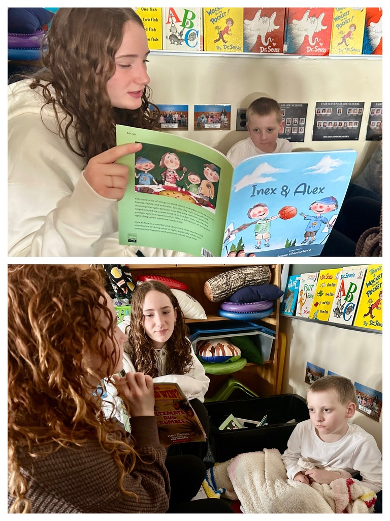 A high school student in a black shirt reads with a young girl holding wooden toys in a classroom filled with books and materials.