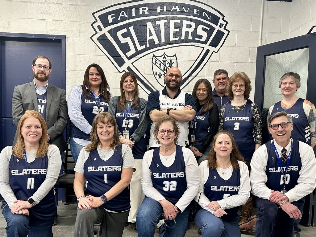 A group of school administrators, faculty, and staff pose together indoors wearing navy “Slaters” basketball jerseys, standing and sitting in front of a wall with a large “Fair Haven Slaters” logo, showing support for the new Unified Basketball Team.
