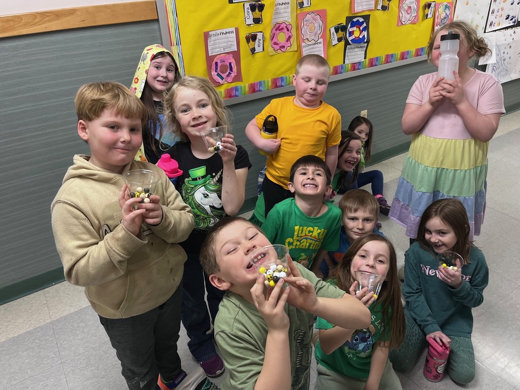 A candid indoor group photograph features nine smiling young children gathered in a school hallway, posing with plastic cups containing black and yellow treats. The child in the foreground, wearing a green t-shirt, holds their cup up and smiles. To their left, a boy in a tan hoodie holds his cup with both hands, while behind him, a girl wearing a black t-shirt with a unicorn design holds hers. On the right, several other children hold their cups, and one girl stands, holding a water bottle up. The children display joyful expressions. They are situated in front of a yellow bulletin board adorned with various school related items, including illustrations of doughnuts and several small, decorative items. The background shows neutral grey and white tiled walls and floors, with a pink plastic water bottle placed on the floor to the right. The lighting is functional for the indoor school setting.