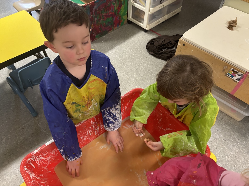 Two preschool students in colorful smocks reach into a red sensory bin filled with orange oobleck, observing and feeling the mixture with their hands.