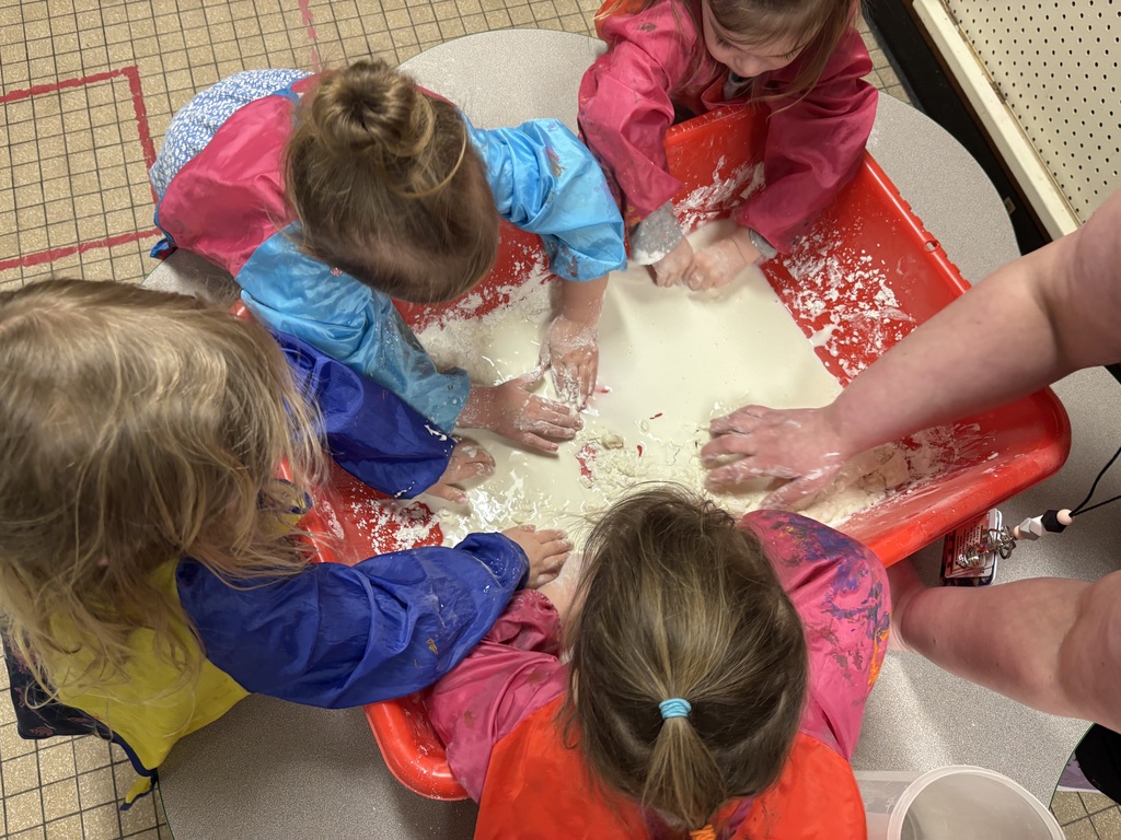 Preschool students in smocks gather around a red sensory bin, exploring oobleck with their hands while a teacher guides the activity.