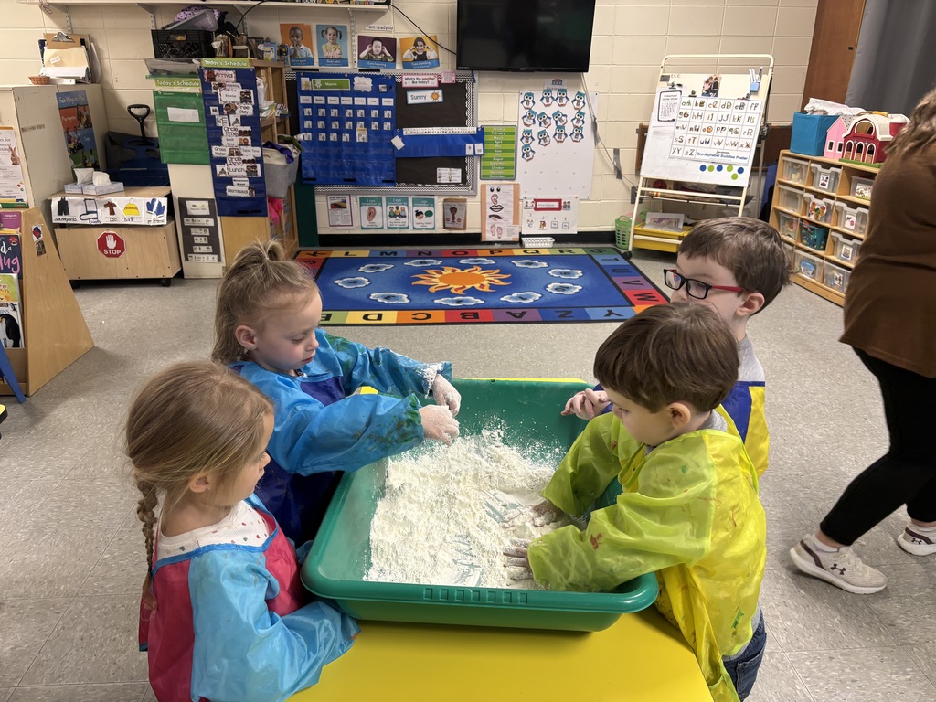 Four preschool students wearing smocks stand around a green sensory bin, using their hands to explore and mix a flour-like substance.