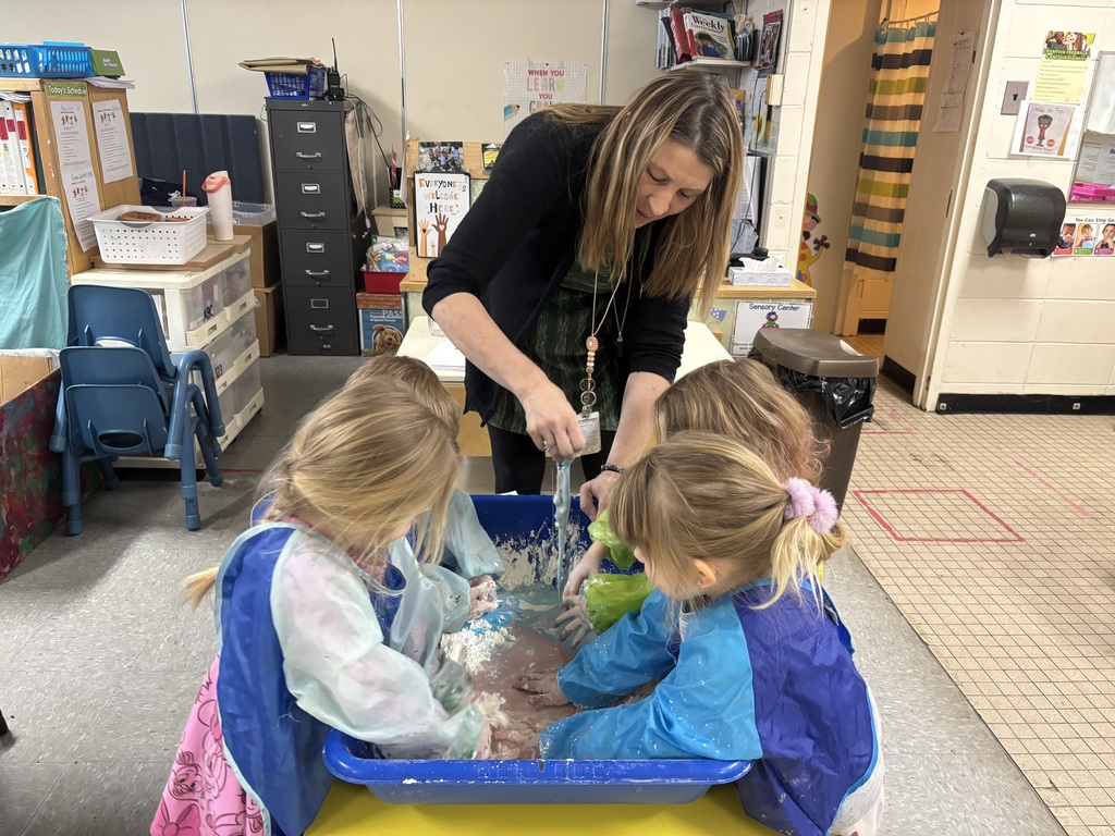 Preschool students wearing colorful smocks gather around a sensory bin, mixing and squeezing oobleck with their hands.
