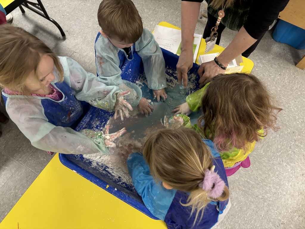 Four preschool students smile and focus on mixing oobleck in a blue sensory bin while a teacher assists nearby.