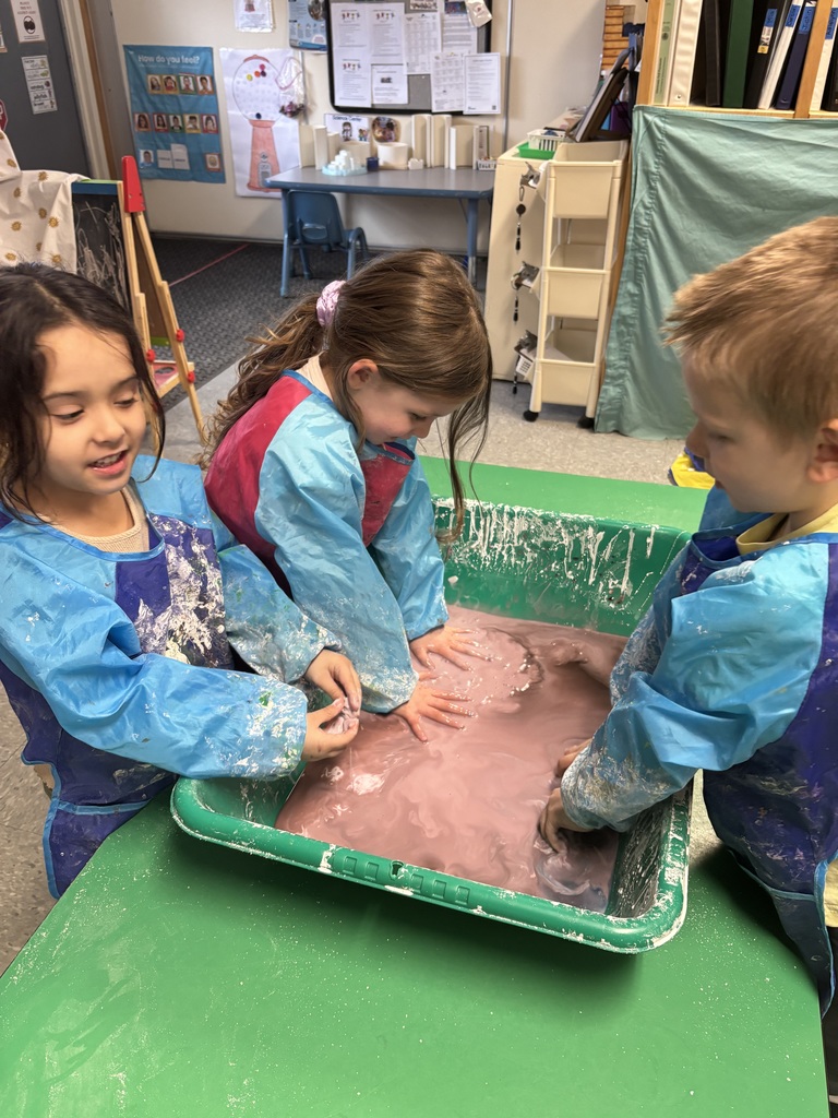 Three preschool students wearing blue smocks stand around a green sensory bin, using their hands to explore and mix pink oobleck in a classroom.