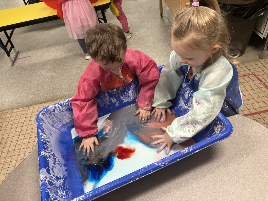Two preschool students wearing smocks press their hands into a blue sensory bin as colorful dye spreads through a milky oobleck mixture, creating swirling patterns.