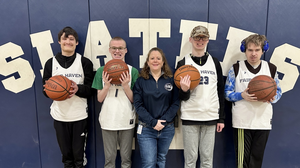 Five individuals stand in a school gym in front of a “Slaters” wall sign. Four high school students wearing Fair Haven basketball jerseys hold basketballs, smiling, while a staff member stands in the center. One student wears earmuffs, and another wears a camouflage hat.