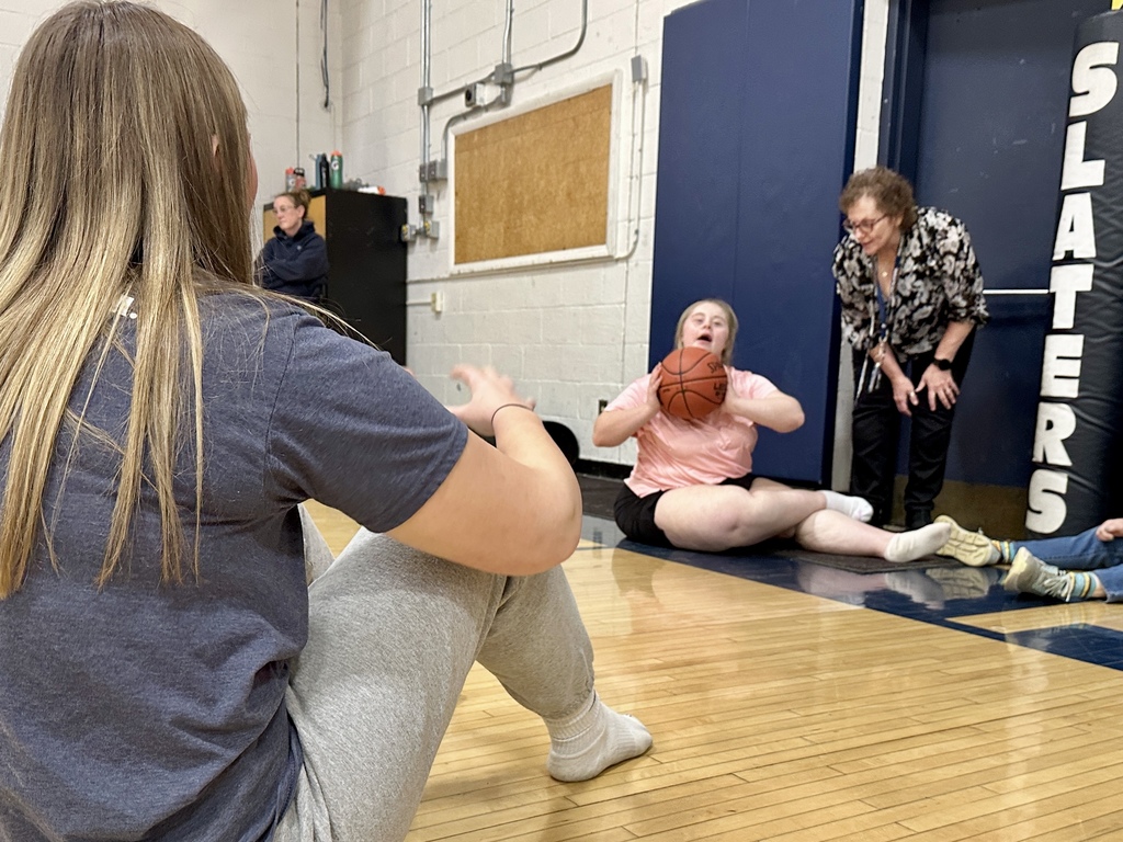 Two students sit on the gym floor passing a basketball back and forth during practice while a staff member leans in to support and encourage them. Another student sits nearby participating in the activity.