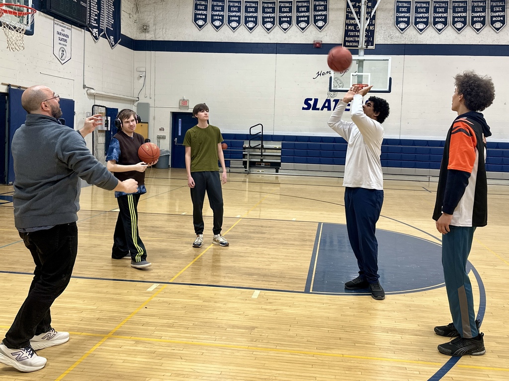 Students and a coach practice basketball in a high school gym. One student takes a shot toward the hoop while others watch, including a coach standing nearby offering guidance. Banners hang on the wall above the court.