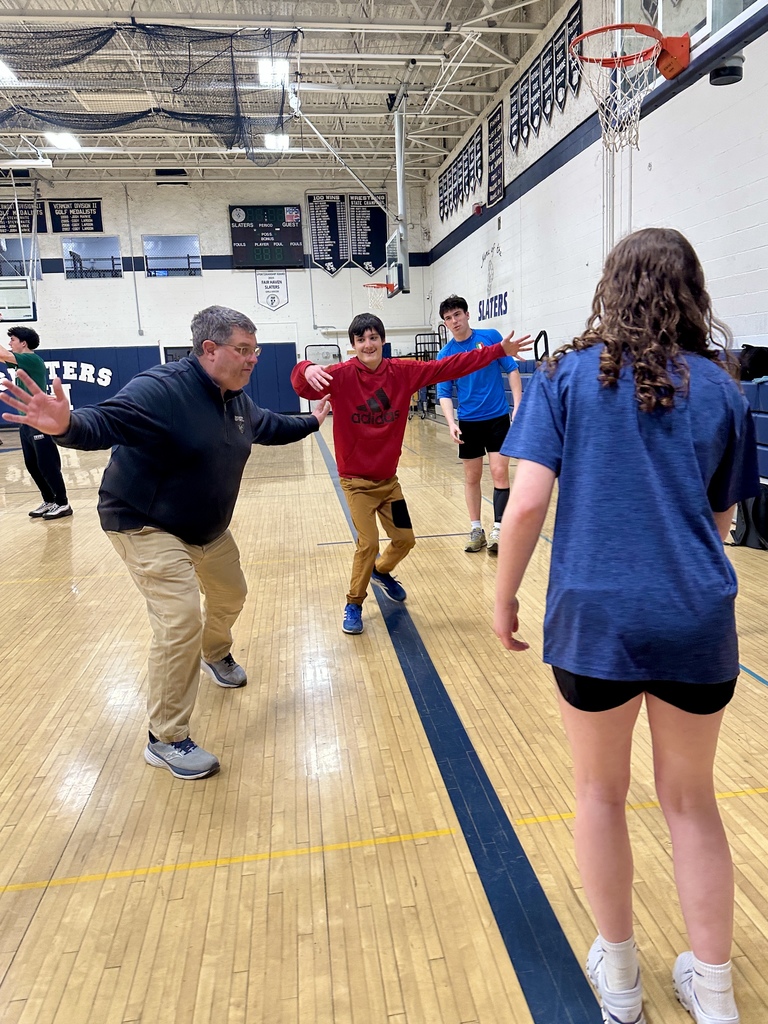 A group of students and a staff member participate in a defensive drill during basketball practice in a gym. One student in a red sweatshirt spreads their arms wide while others move around the court.