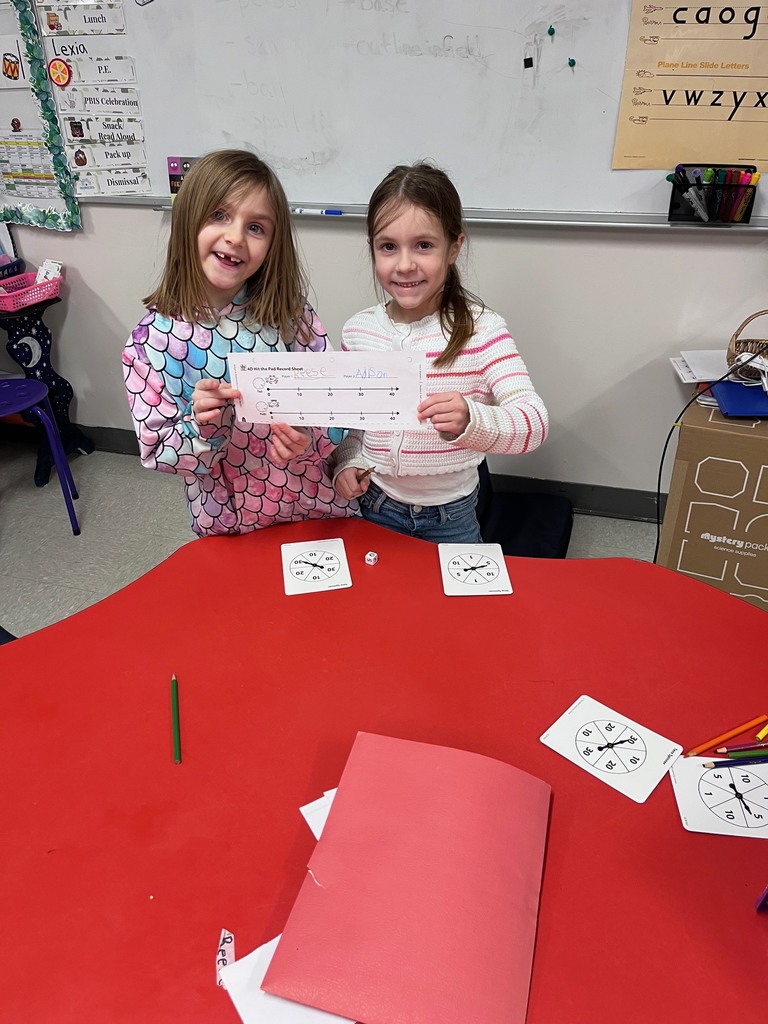 Two girls stand behind a red table, proudly holding up a math worksheet featuring two number lines. On the table are several circular spinners used for their math game.