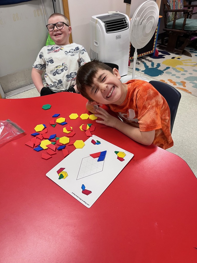 mage 5: Two boys sit at a red table, laughing joyfully. In front of them are a pile of colorful plastic pattern blocks and a white game board featuring a diamond-shaped grid.