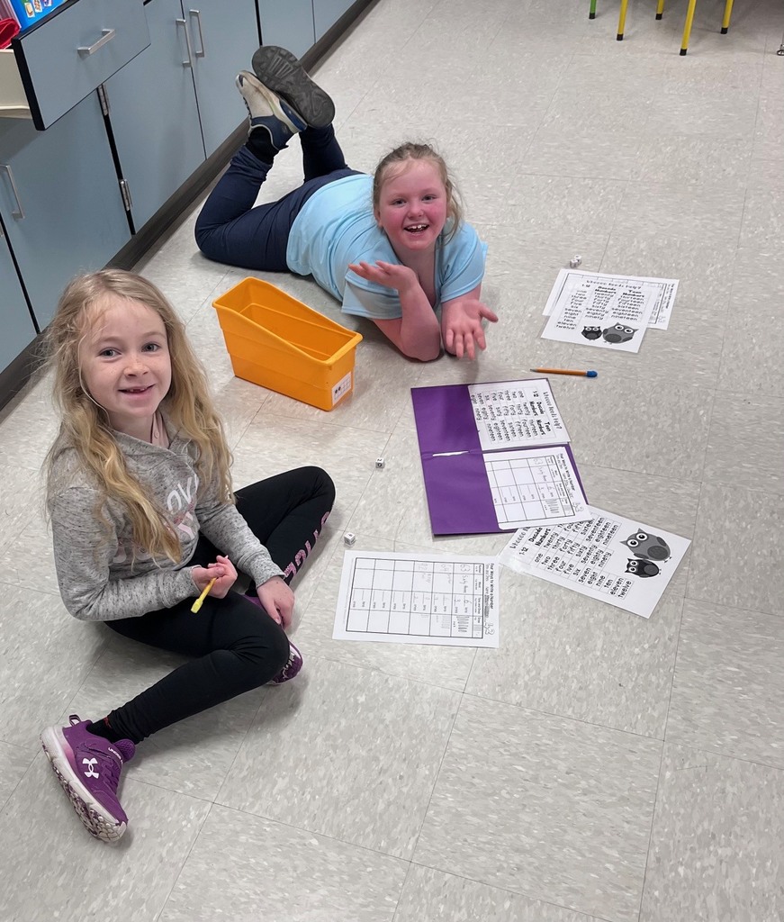 An overhead medium-long photograph captures two young girls playing a game on the classroom floor. On the left, a girl with long blonde hair, wearing a gray sweatshirt and black leggings, sits cross-legged. She holds a yellow marker and smiles towards the camera. Next to her is a second girl with curly brown hair, wearing a light blue t-shirt and dark pants, who is lying on her stomach with her legs kicked back in the air. She smiles broadly with her hands outstretched. They are surrounded by various game materials: a yellow bin, dice, pencils, an open purple folder containing a worksheet, and other loose worksheets on the floor. In the background are light-blue cabinets with metal handles and part of a white table leg. The lighting is bright and even.
