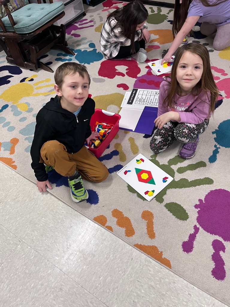 A boy and a girl sit on a colorful rug next to a red bin filled with pattern blocks. They are working together to fill a diamond-shaped outline on a game board with green, red, and yellow shapes.