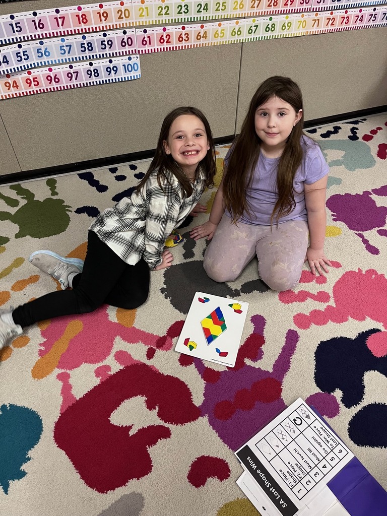 Pattern Blocks & Number Lines Image 1 & 1 (1): Two young girls smile while sitting on a colorful "paint splatter" rug. Between them is a game board titled "Last Shape Wins," which they have partially filled with colorful geometric pattern blocks like hexagons and trapezoids.