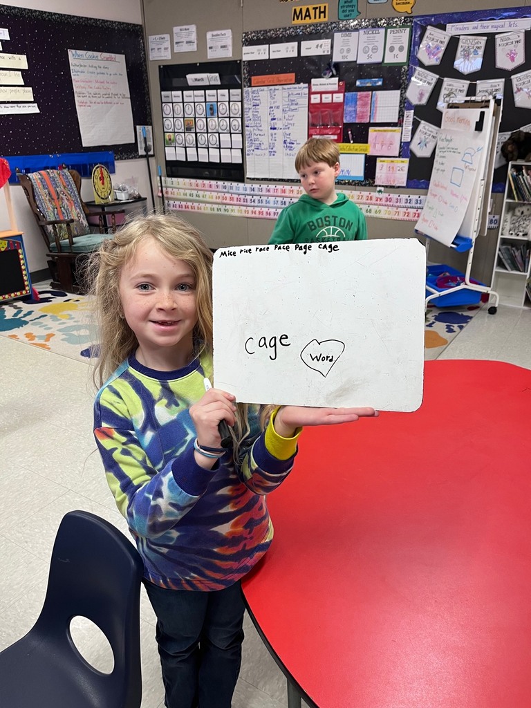 A young girl with blonde hair smiles as she holds up a small whiteboard. On the board, she has written the word "cage" and drawn a heart with the word "word" inside it.