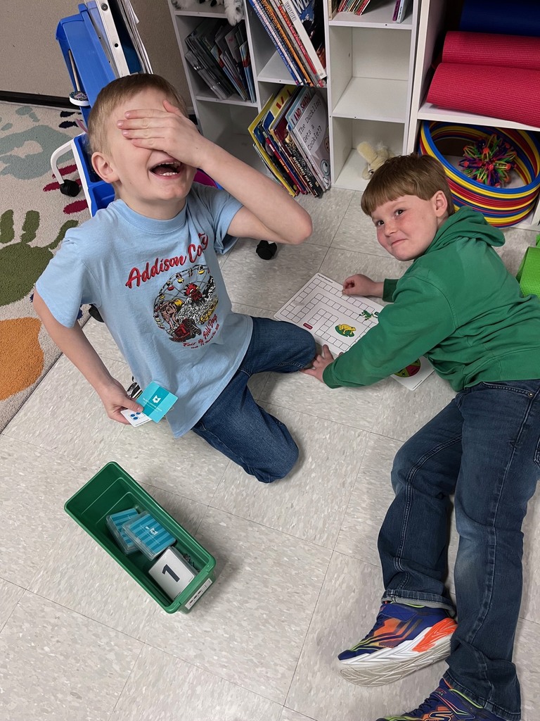 A medium-long high-angle photograph showing two young boys engaged with a board game on a classroom floor. A boy on the left, with blonde hair, is kneeling on the tiled floor and laughing, covering his eyes with his right hand. He is wearing a light-blue t-shirt and blue jeans and holds game cards in his left hand. The t-shirt features a red logo and white graphic of children around a large wheel with text: "Addison County, For all the kids of the world, for all the world's kids." To his right, a second boy lies on his back on the floor, propped up slightly, looking towards the camera. He has short auburn hair and is wearing a green hoodie, blue jeans, and blue and orange sneakers with purple laces. In front of him, on the floor, is a small square whiteboard with a green-drawn grid and drawings of two frogs on the left. A green plastic box lies on the floor between the boys, containing two white square blocks, each with the number "1" written in black. The background shows a wooden bookshelf with books and red and blue plastic rolling tubes and multi-colored hoops on wheels. Part of a small blue table leg is visible on the upper left. The tiled floor is a speckled light gray.
