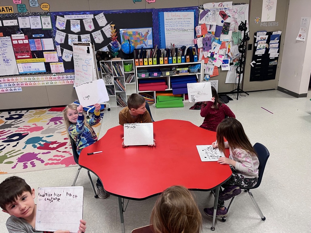 Four children sit at a red table in a classroom, showing off their whiteboards. The boards contain lists of words ending in "-ice" and "-age." A colorful bulletin board with a map and student work is visible in the background.