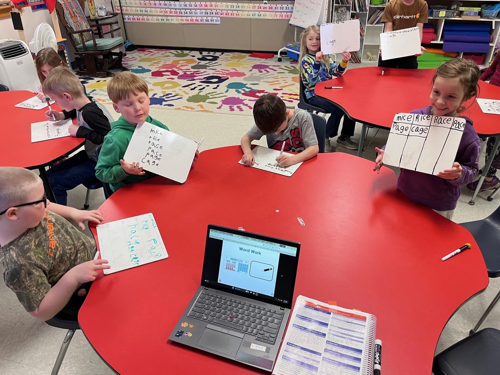 A classroom scene where students at a red table are engaged in "Word Work." One girl holds up a whiteboard with words organized into columns, while a laptop in the foreground displays a digital lesson on the same topic.