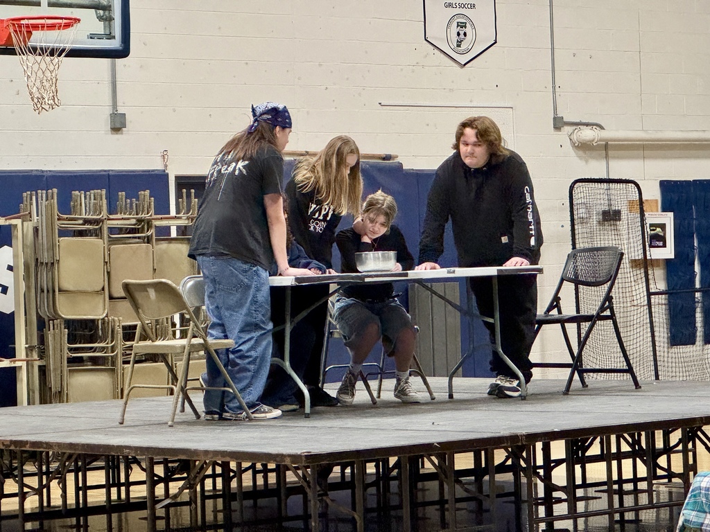 Four students stand around a table on a stage in a gym, acting out a scene during a drama performance rehearsal.