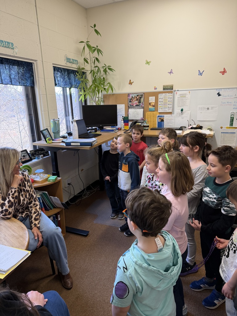 A group of first grade students stands in a classroom gathered around a seated teacher, smiling and singing to her during a birthday celebration.