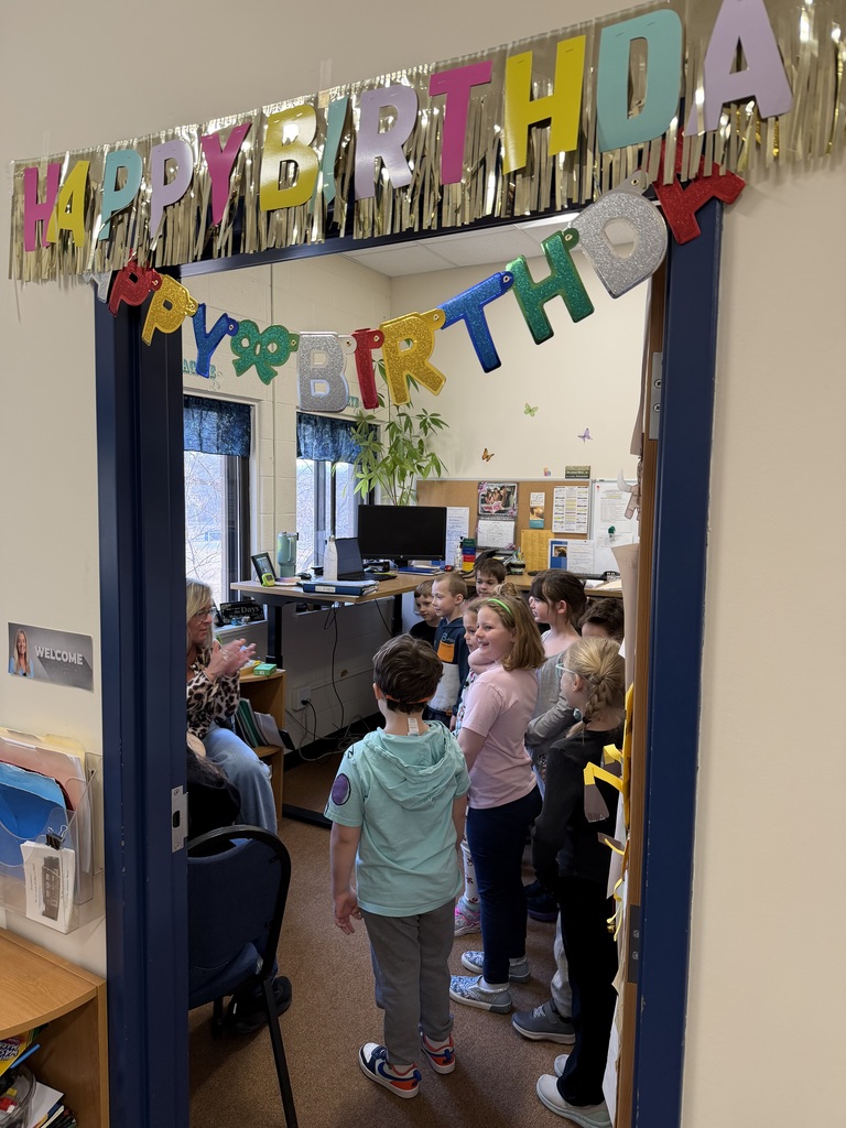 A classroom doorway decorated with colorful “Happy Birthday” banners frames a group of young students singing to their teacher inside the room.
