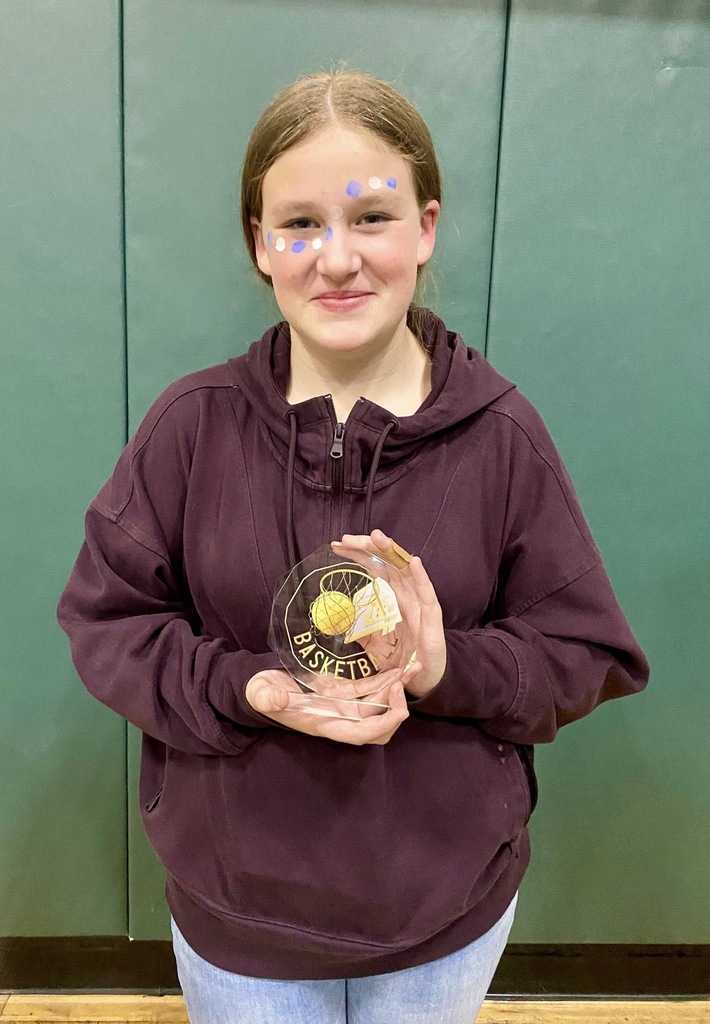 A student stands in a gym holding a clear basketball award, smiling with decorative face paint, against a green padded wall.