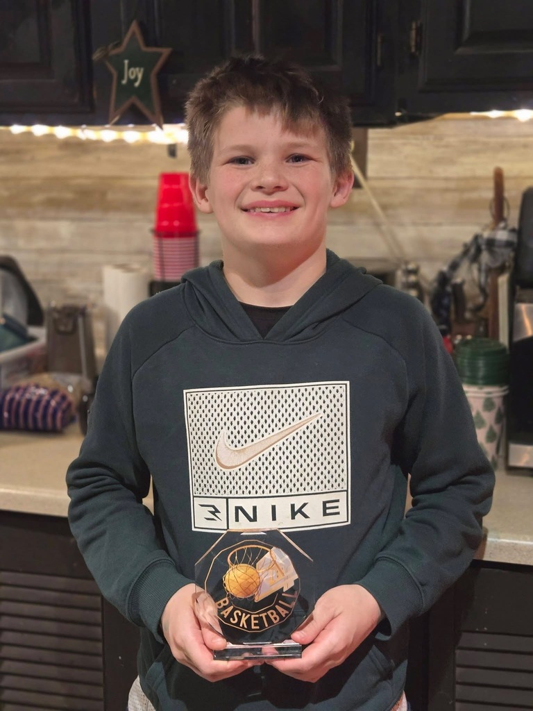 A young boy stands in a kitchen holding a basketball award, smiling proudly while wearing a dark green sweatshirt.