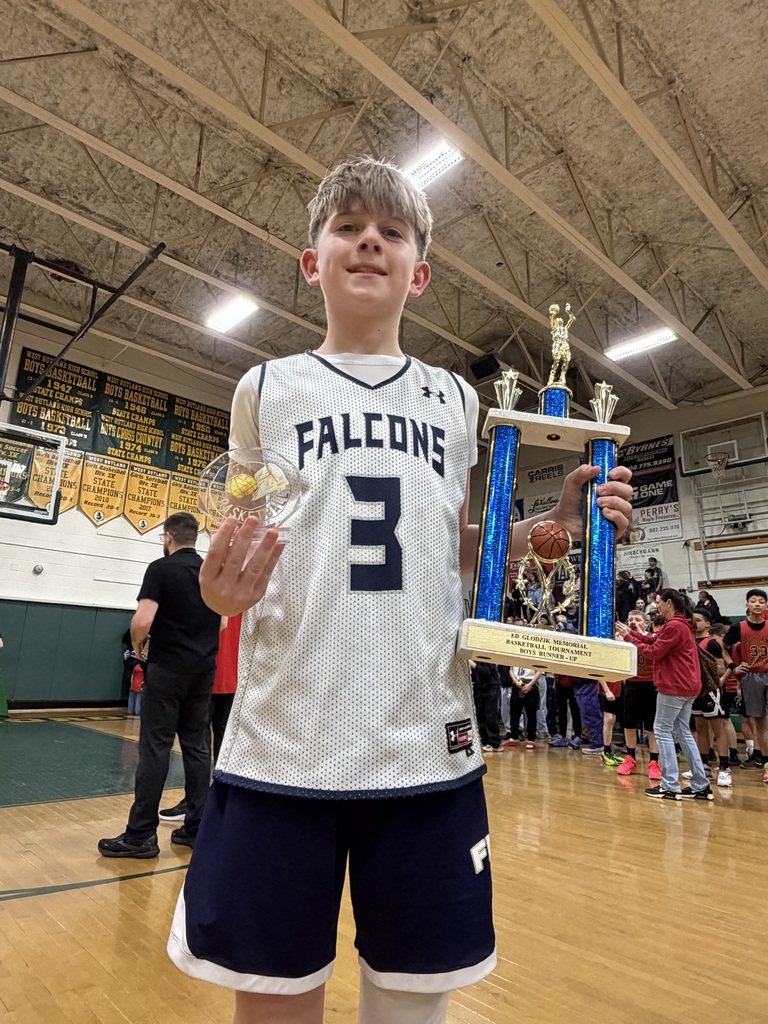 A young basketball player in a Falcons jersey holds both a trophy and a small award on a gym court with other players and spectators in the background.