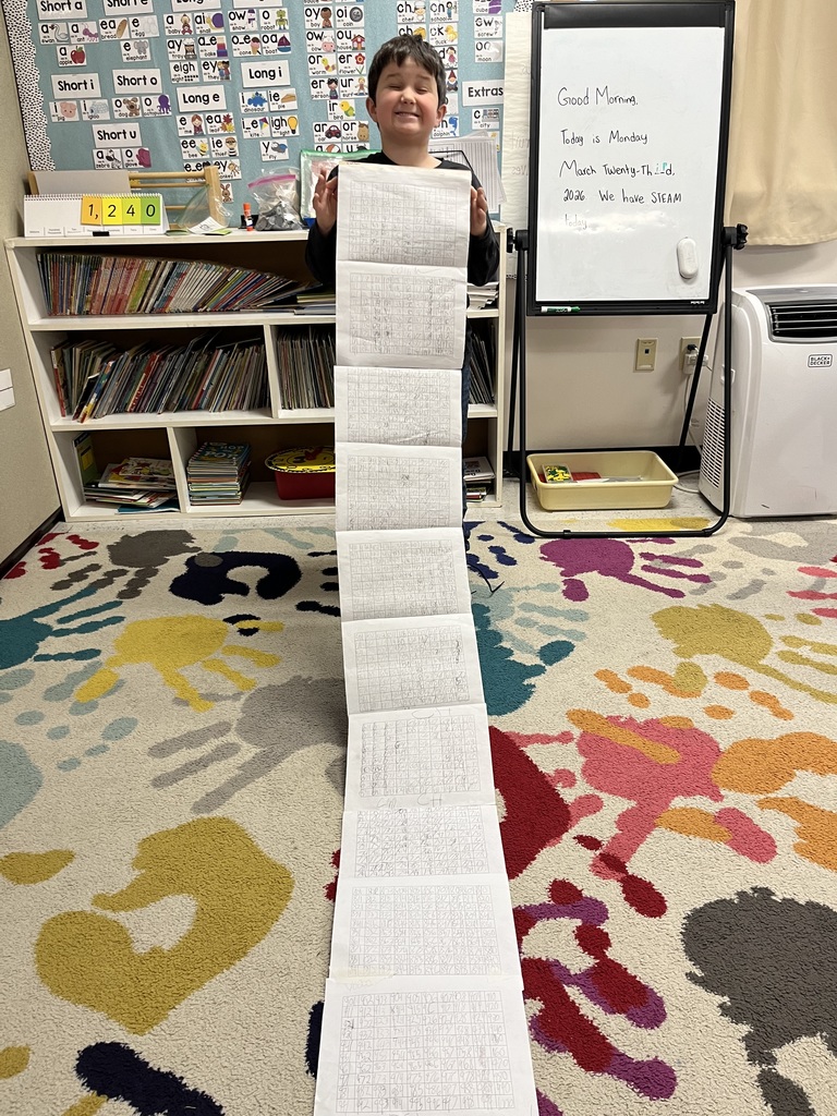 A young boy with a joyful expression holds up a long paper scroll filled with hand-written numbers. He is standing on a vibrant handprint-patterned rug next to a bookshelf and a whiteboard dated March 23, 2026.IMG_4925.jpgA smiling young boy wearing a "Miles Morales" shirt holds a long, rolled-out scroll of number grids. The scroll extends down to the classroom floor, where another student is visible in the background.