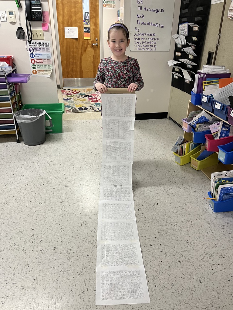 A young girl with a purple headband smiles proudly while holding the top of a long paper scroll. The scroll is filled with mathematical grids and numbers, extending down to a light-colored tile floor.IMG_4919.jpgA young girl with long brown hair stands in a classroom doorway, holding up a long, hand-written number scroll that unfurls onto the floor. In the background, a whiteboard shows a schedule and student initials.