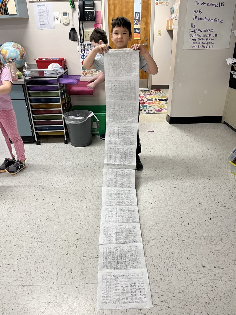 A young boy holds the top of a very long, multi-page paper scroll covered in rows of numbers. The scroll reaches the floor and extends forward, showcasing his hard work in a bright classroom.