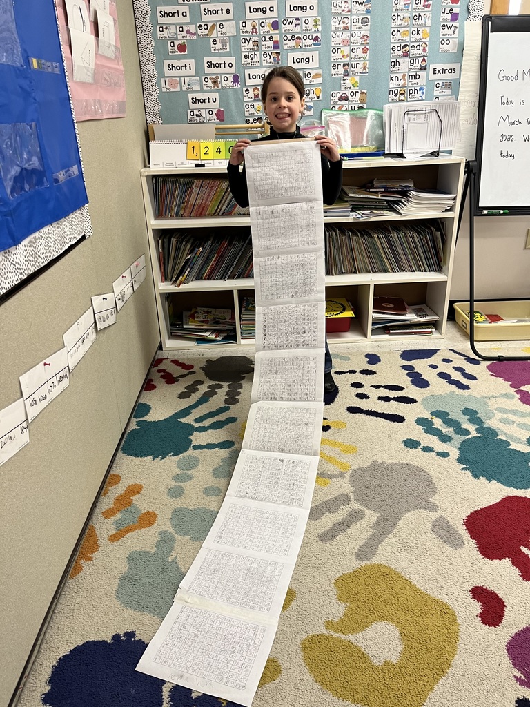 A young girl smiles while holding a long scroll of number charts in the corner of a classroom. She is standing on a colorful rug featuring large, multi-colored handprints
