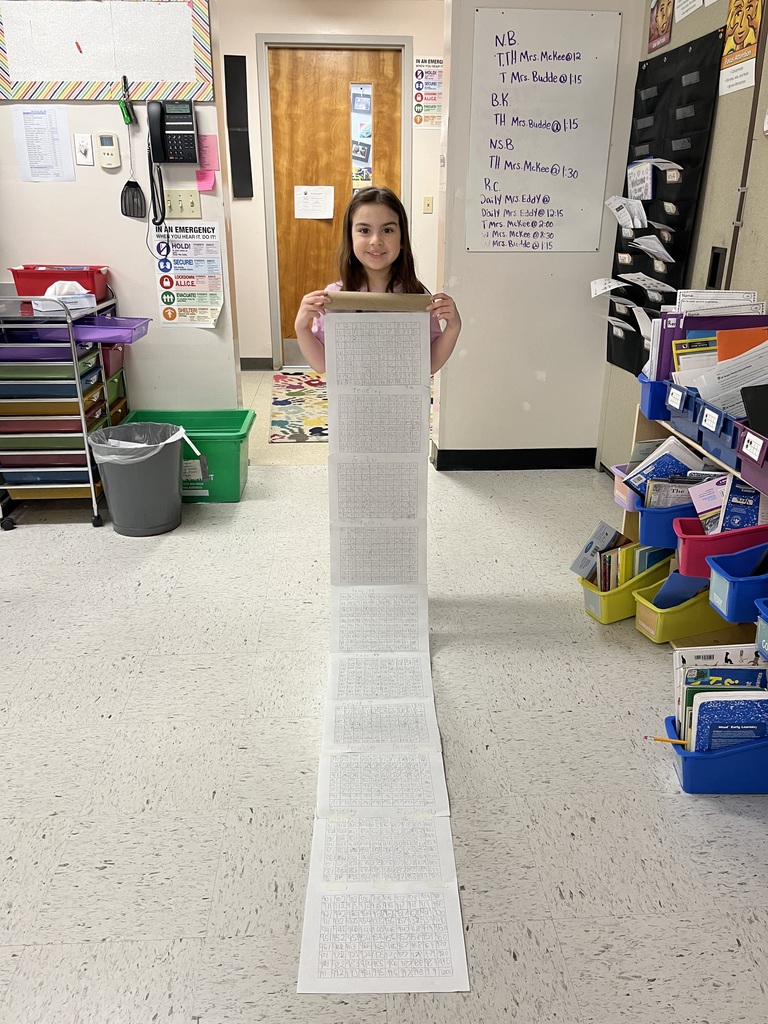 A young girl with long brown hair stands in a classroom doorway, holding up a long, hand-written number scroll that unfurls onto the floor. In the background, a whiteboard shows a schedule and student initials.IMG_4921.jpgA young boy holds the top of a very long, multi-page paper scroll covered in rows of numbers. The scroll reaches the floor and extends forward, showcasing his hard work in a bright classroom