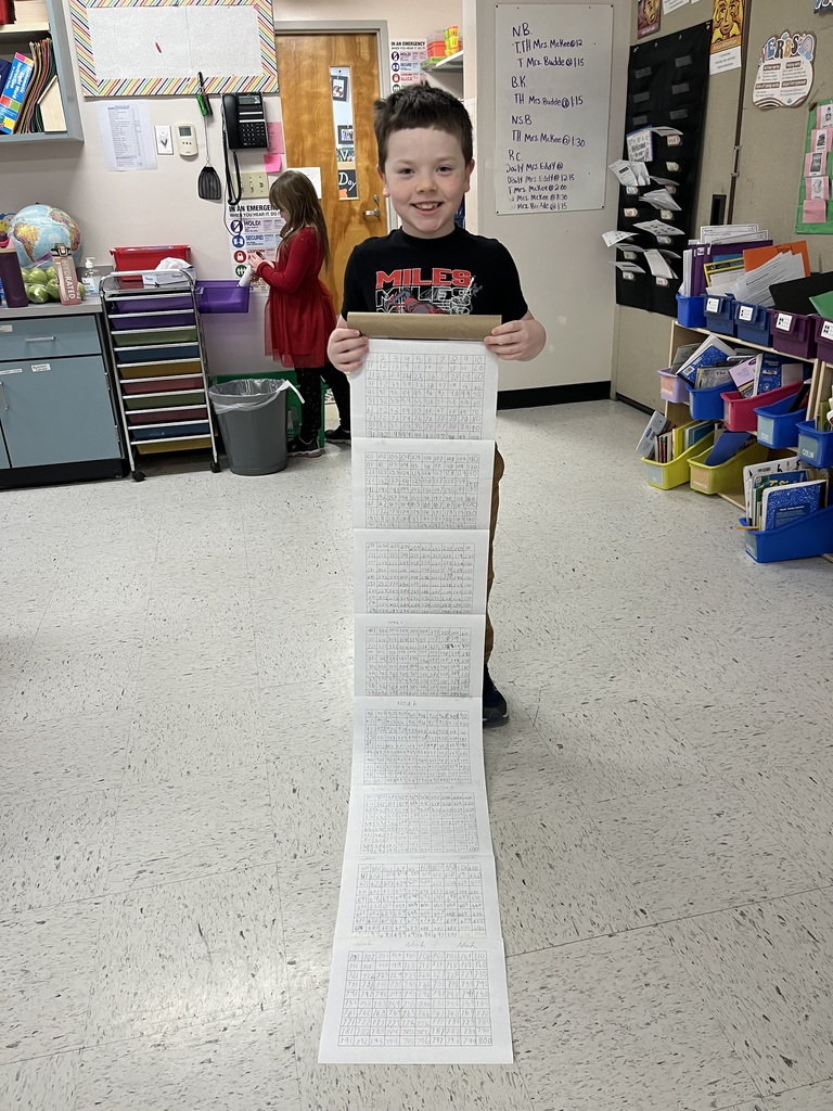 A smiling young boy wearing a "Miles Morales" shirt holds a long, rolled-out scroll of number grids. The scroll extends down to the classroom floor, where another student is visible in the background.