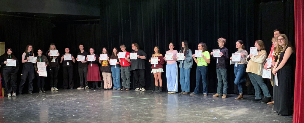 A group of high school students stand on a stage holding certificates, smiling and posing together after a drama festival performance.