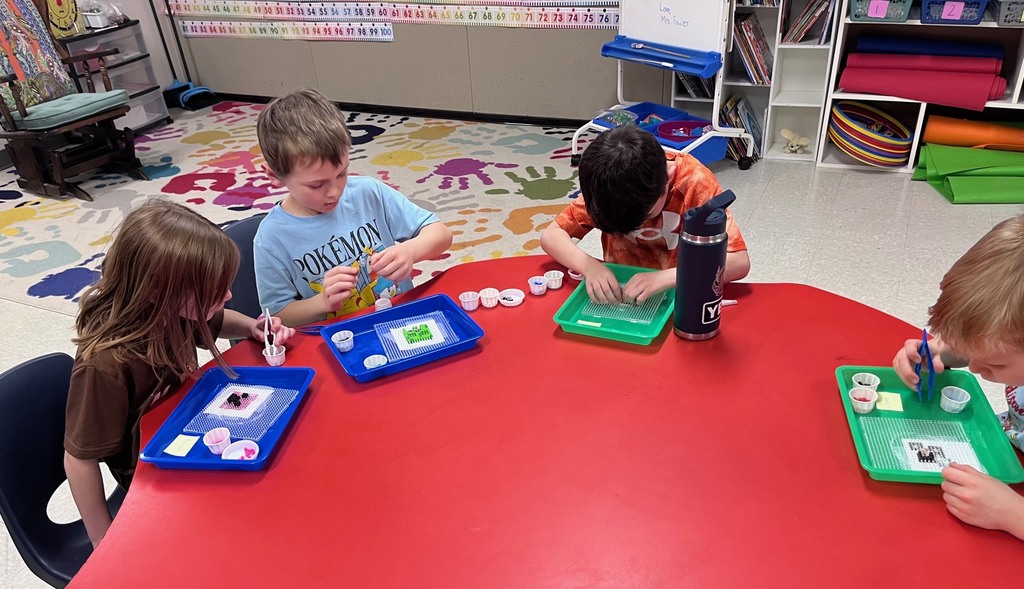 Text A group of four elementary-aged children sit around a large, red clover-shaped table in a classroom. They are focused on a craft project using Perler beads (meltable plastic beads), using small plastic tweezers to arrange beads onto pegboards set inside individual colorful plastic trays. One boy wears a light blue Pokémon shirt, and a large black Yeti water bottle sits on the table. The background shows a classroom environment with a colorful handprint rug, a number line, and bookshelves. Photo 2 Alt Text High-angle shot of seven children gathered around a large, red activity table in a classroom, working on bead art. Each child has a colorful tray (pink, yellow, blue, or red) containing a pegboard and small cups filled with various colored beads. Some children are using their fingers while others use blue plastic tweezers to carefully place the beads. The children are dressed in casual school clothes, including a green "Boston" hoodie and a light blue t-shirt.