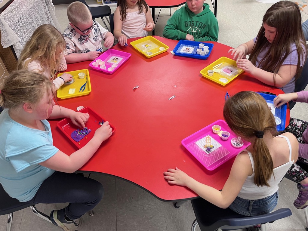 Text High-angle shot of seven children gathered around a large, red activity table in a classroom, working on bead art. Each child has a colorful tray (pink, yellow, blue, or red) containing a pegboard and small cups filled with various colored beads. Some children are using their fingers while others use blue plastic tweezers to carefully place the beads. The children are dressed in casual school clothes, including a green "Boston" hoodie and a light blue t-shirt.