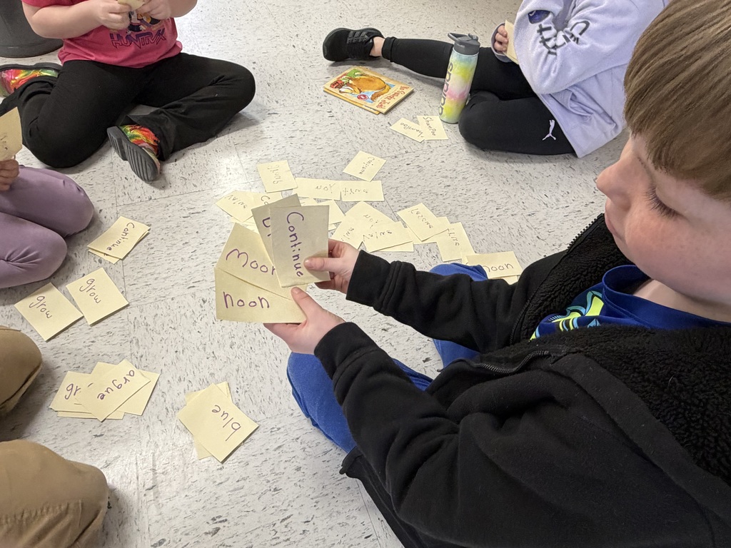 A close-up, high-angle shot of a young boy in a black hoodie holding a fan of yellow word cards. The card on top clearly shows the word "Continue." Other cards on the floor show "Moon," "noon," and "blue."
