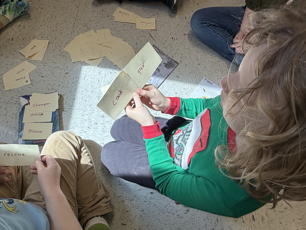 A close-up focus on a child’s hands holding two yellow slips of paper labeled "cook" and "grew." In the background, other slips of paper with words like "shampoo," "moon," and "rescue" are scattered on the floor.