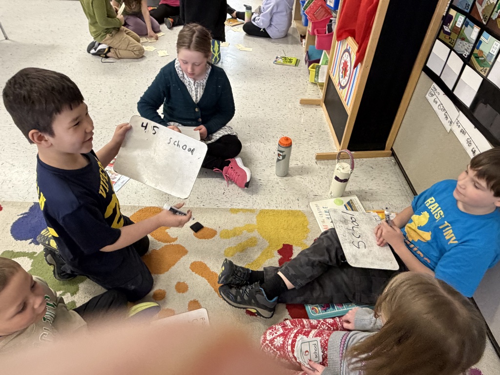 A group of students sit on a colorful paw-print rug. Two boys hold up small whiteboards; one has the number "45" and the word "school" written on it. They appear to be practicing spelling or vocabulary in a small group. IMG_0284.jpg: Five children sit on a patterned rug working with whiteboards and markers. One girl in the foreground writes the words "So," "Sound," and "+100" on her board. Educational posters and a bookshelf are visible in the background.
