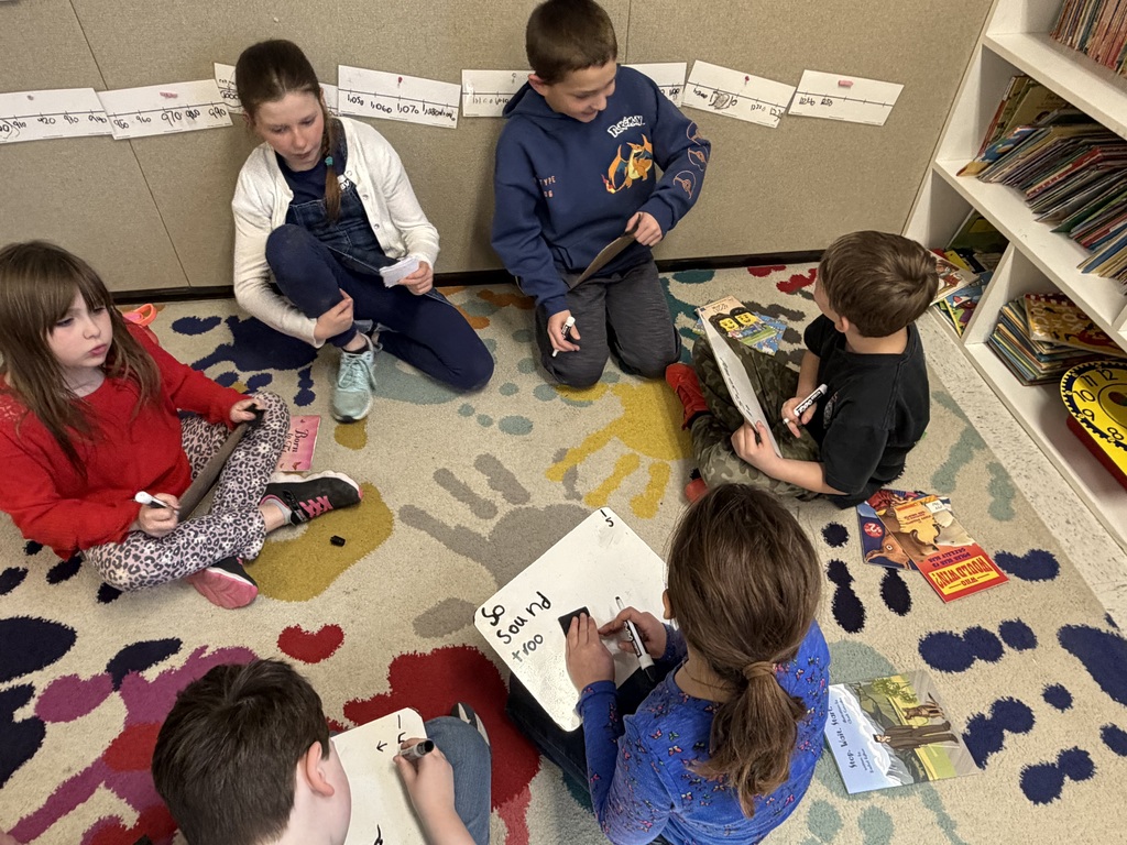 : Five children sit on a patterned rug working with whiteboards and markers. One girl in the foreground writes the words "So," "Sound," and "+100" on her board. Educational posters and a bookshelf are visible in the background.