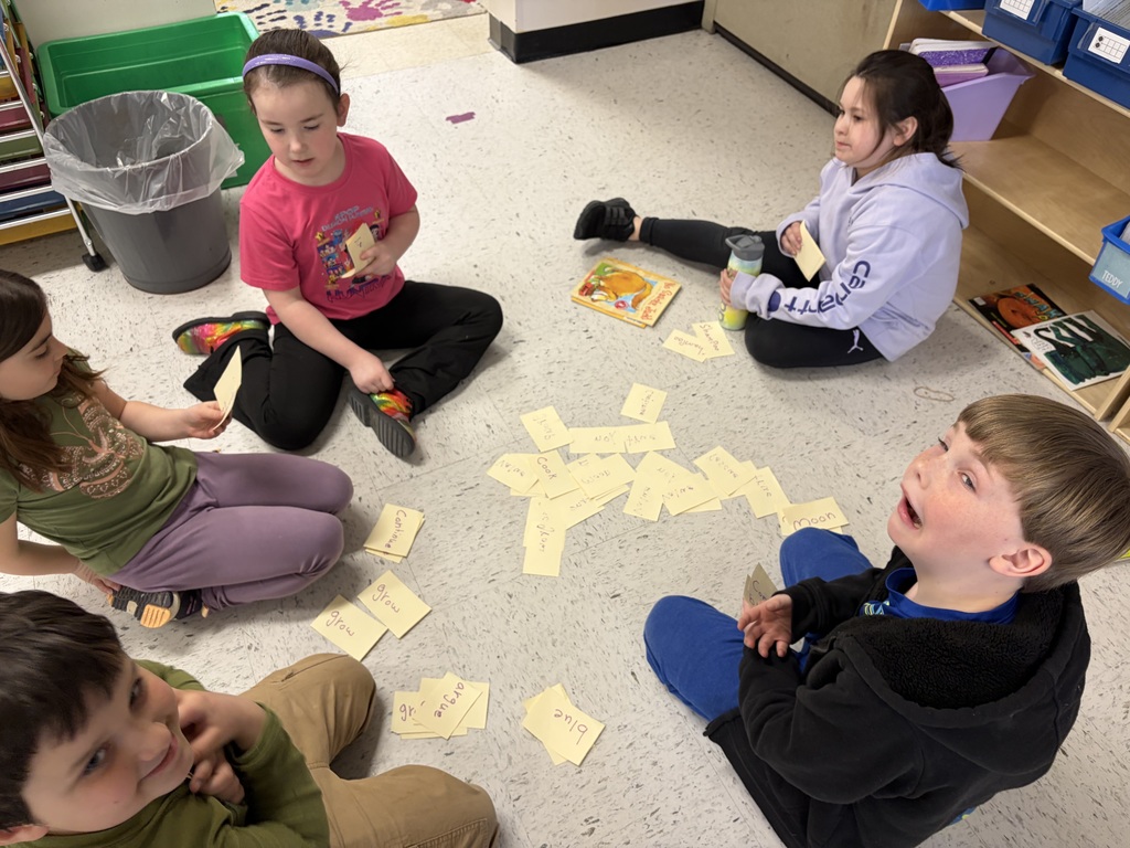 Five children sit in a circle on a classroom floor, surrounded by small yellow slips of paper with handwritten words. They are engaged in a word-sorting or matching game.