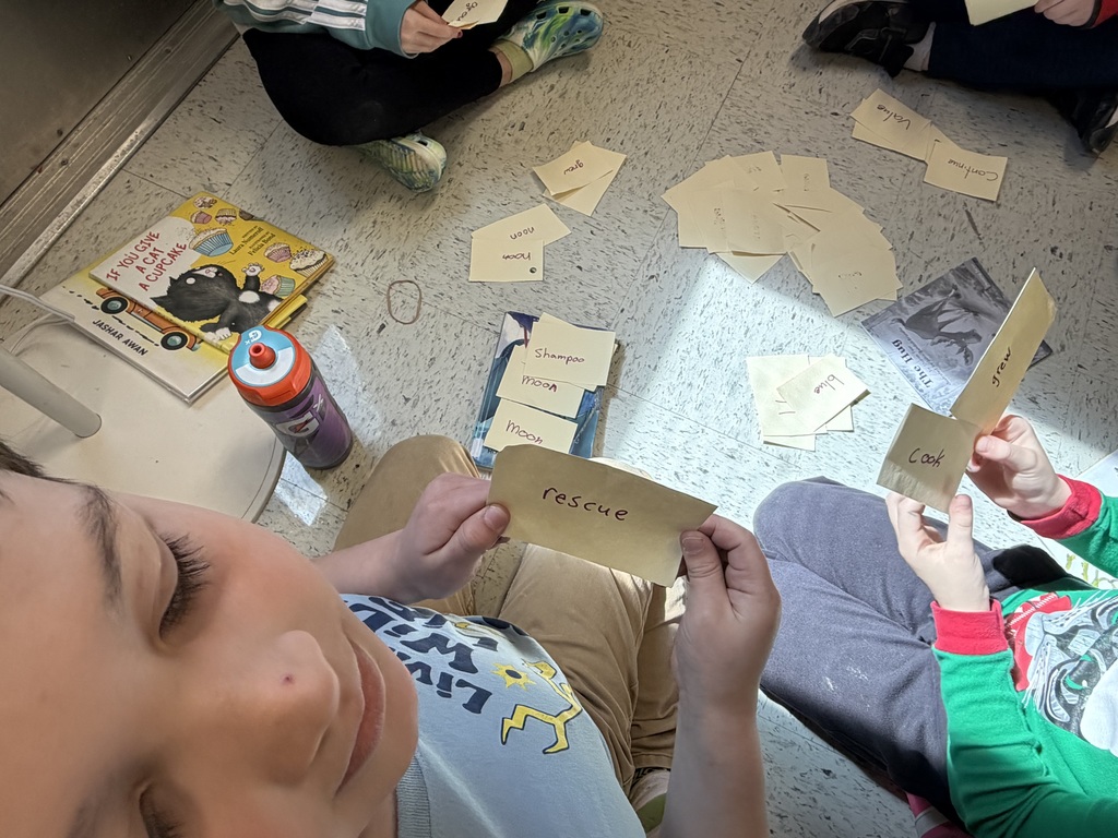 A top-down view of two children holding yellow word cards. One child holds a card that says "rescue," while the other holds cards saying "cook" and "grew." Several books, including If You Give a Cat a Cupcake, lie on the floor nearby. IMG_0286.jpg: A close-up focus on a child’s hands holding two yellow slips of paper labeled "cook" and "grew." In the background, other slips of paper with words like "shampoo," "moon," and "rescue" are scattered on the floor.