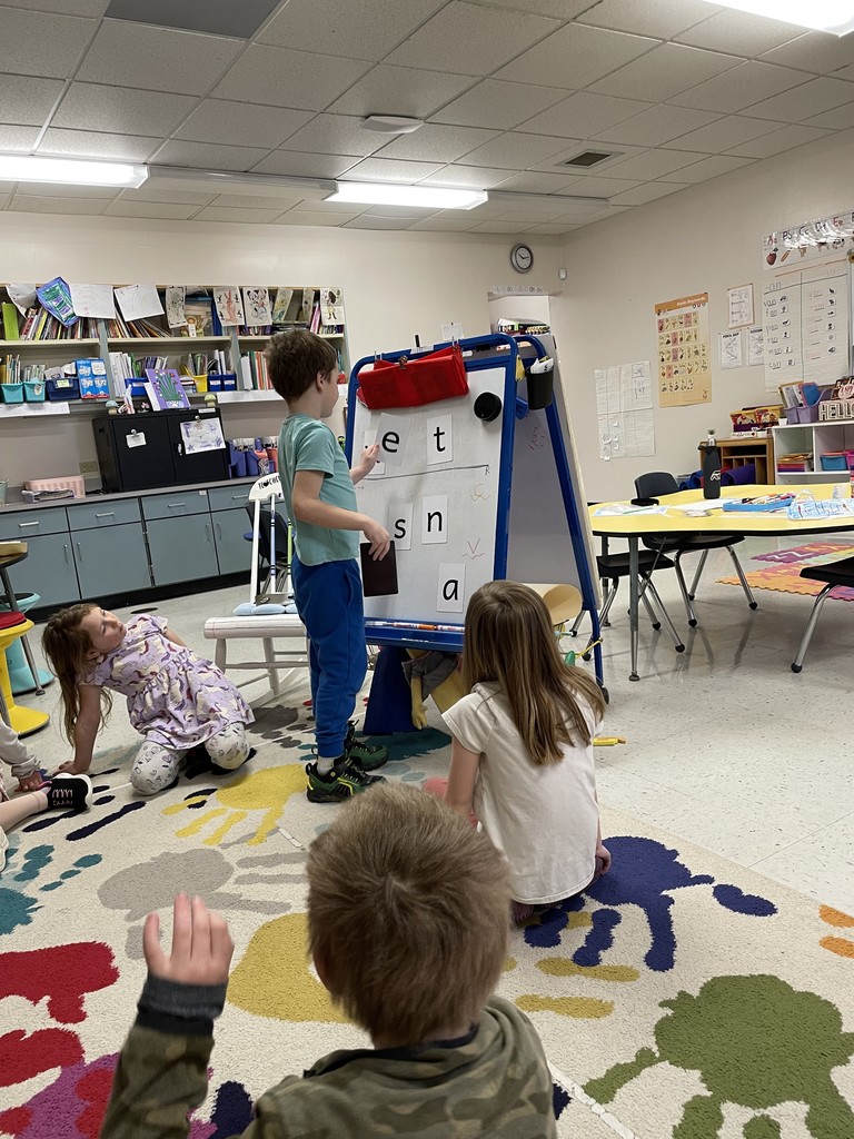 A young boy in a teal t-shirt and blue joggers stands at the whiteboard, pointing to the letter "e" next to a "t." He is leading a literacy activity for a small group of classmates who are sitting on a large, patterned rug. The wide-angle shot shows the classroom environment, including a yellow activity table, educational posters on the walls, and fluorescent ceiling lights. Image 4: Boy in Camo Hoodie
