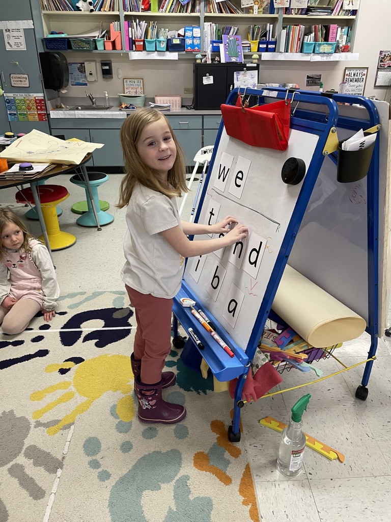 A young girl with blonde hair, wearing a white t-shirt, pink pants, and purple rain boots, stands at a blue mobile whiteboard easel in a colorful classroom. She is smiling at the camera while arranging large letter cards on the board. The letters "w," "e," "t," "n," "d," "i," "b," and "a" are visible. Another student sits on a colorful rug in the background. Image 2: Girl in Blue Long-Sleeve