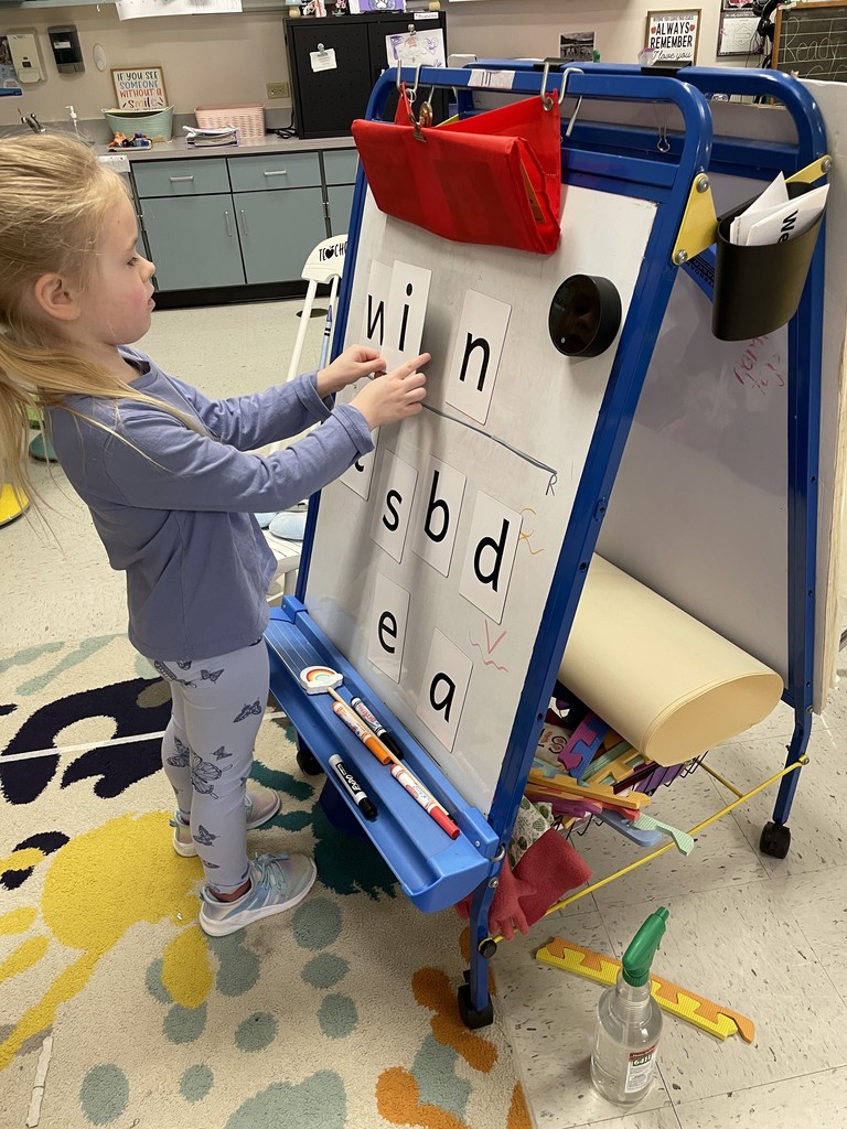 A young girl with a blonde ponytail, wearing a blue long-sleeve shirt and butterfly-patterned leggings, focuses on a whiteboard easel. She is using both hands to position the letter cards "w," "i," and "n" in a row. Below them, other letters like "s," "b," "d," "e," and "a" are scattered. The classroom background shows shelves filled with books and organized bins. Image 3: Boy in Teal Shirt