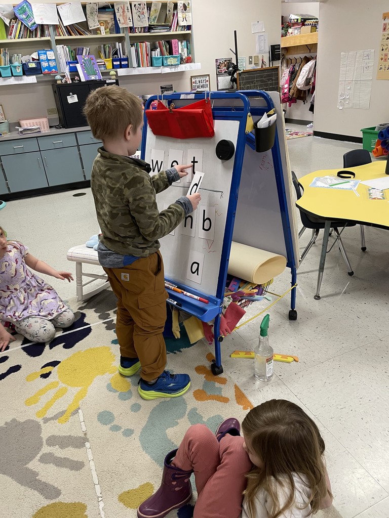 A young boy wearing a camouflage hoodie and tan cargo pants stands at the blue easel. He is holding a letter "d" card and pointing toward the word "wet" spelled out at the top of the board. A girl in a floral dress watches from the rug nearby. Markers and a spray bottle are visible on the easel’s tray and the floor.