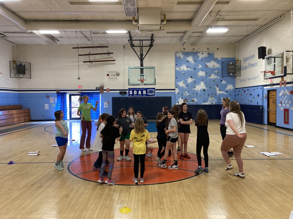 Girls and coaches standing in a circle on a gym court engaging in an active group activity, with cones and equipment visible on the floor.