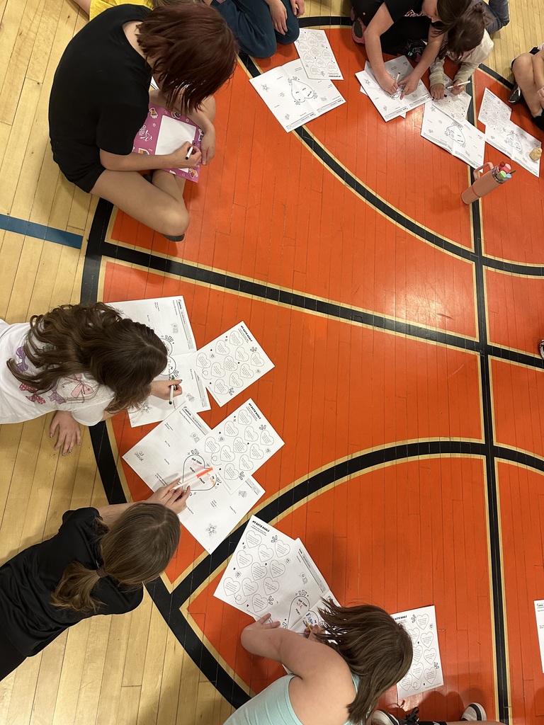 Overhead view of girls sitting on a gym floor working on worksheets with drawings and writing, focusing on activities about identity and personal traits.